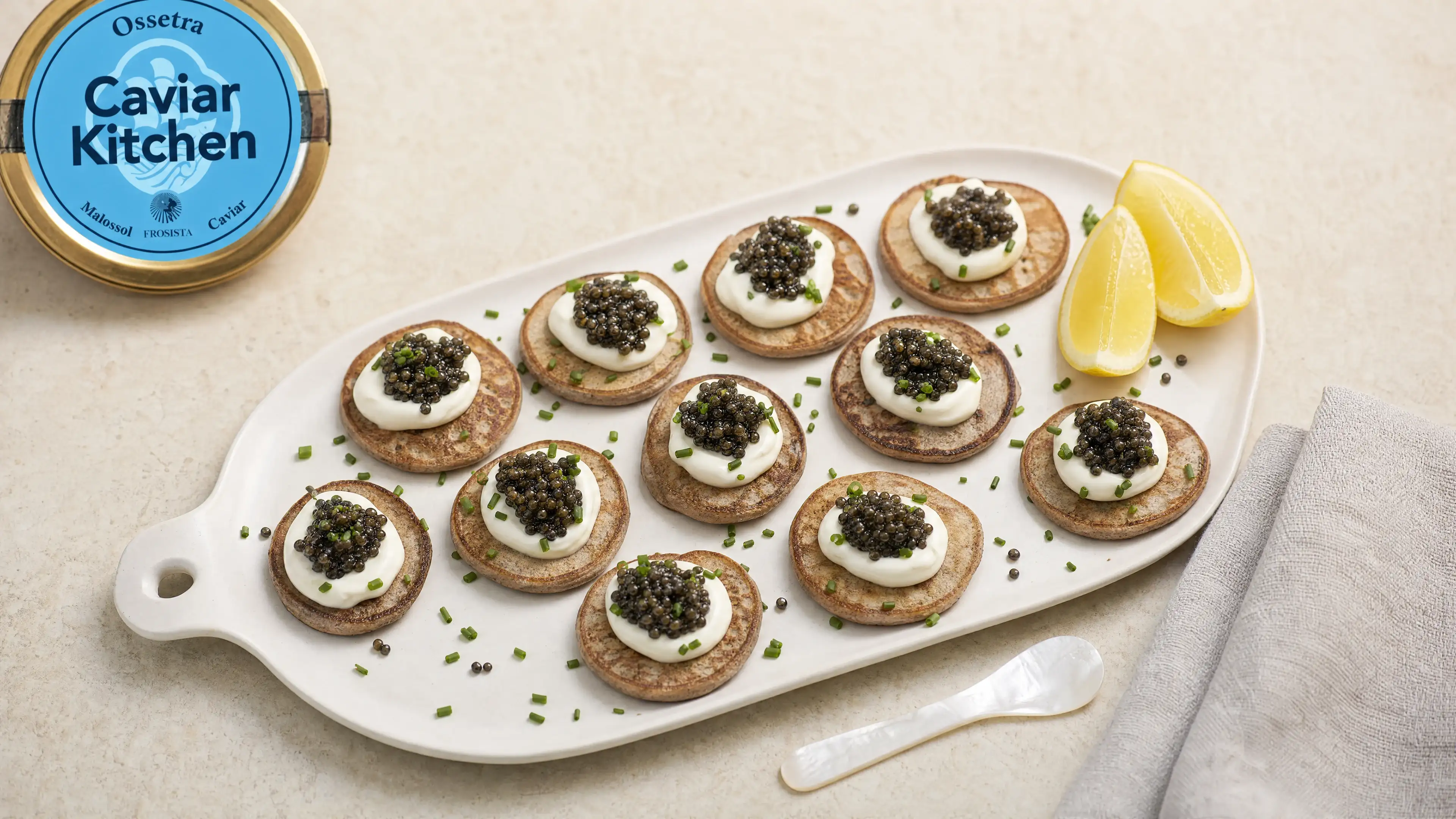 Twelve buckwheat blini topped with crème fraîche and Ossetra caviar on a white ceramic board with lemon wedges and chives