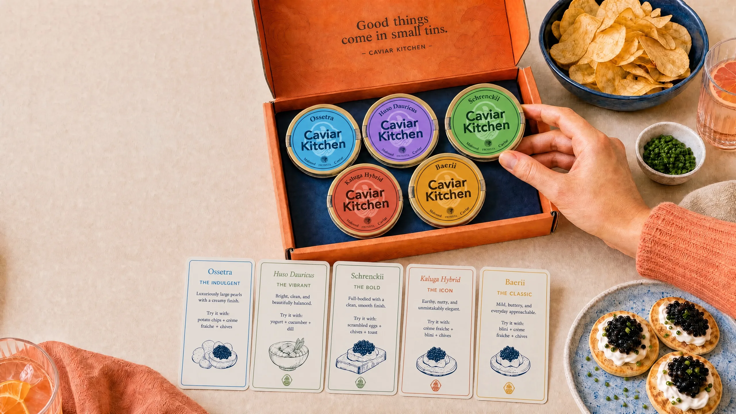 A Caviar Kitchen tasting box of five caviar tins with chips, blini, and drinks on a table.