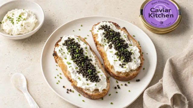 Two slices of sourdough toast spread with whipped goat cheese and topped with dark caviar and chives on a white ceramic plate